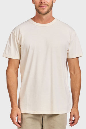 A man wears the Academy Brand Jimmy Tee in plain off-white 100% cotton with beige pants, standing against a light gray background, facing forward with arms relaxed at his sides.