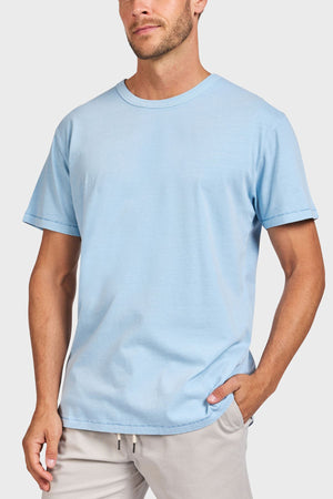 A man wearing the Academy Brand Jimmy Tee in light blue and light beige pants stands with one hand in his pocket against a plain light background.
