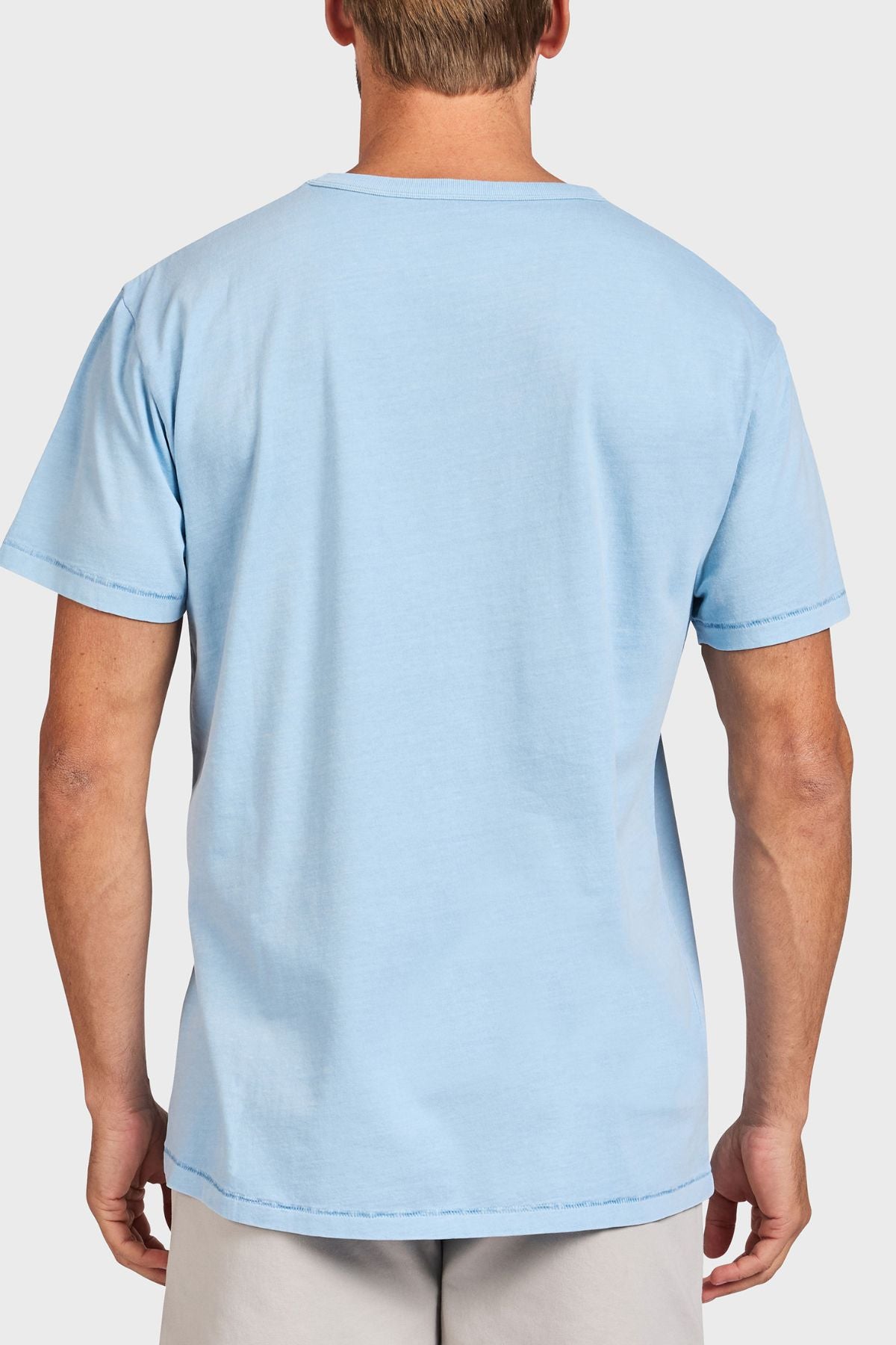A person is seen from the back wearing the Academy Brand Jimmy Tee in light blue with light-colored shorts, set against a plain white background.