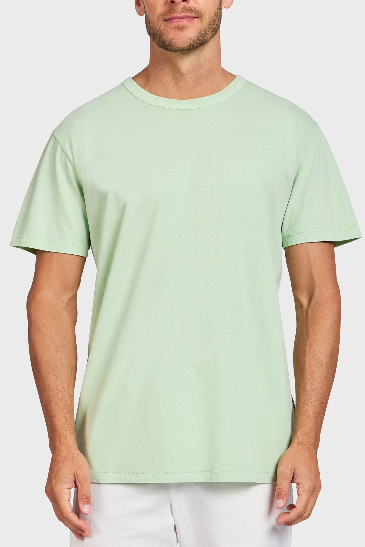 A man wears the Academy Brand Jimmy Tee, a light green loose fit t-shirt made of 100% cotton, paired with white pants. He stands against a plain white background with his arms relaxed at his sides.