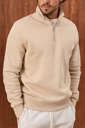 A man wears the Academy Brand 1/4 Zip Sweat with light gray sweatpants, standing hands-in-pockets against a wooden background. His face is partly visible.