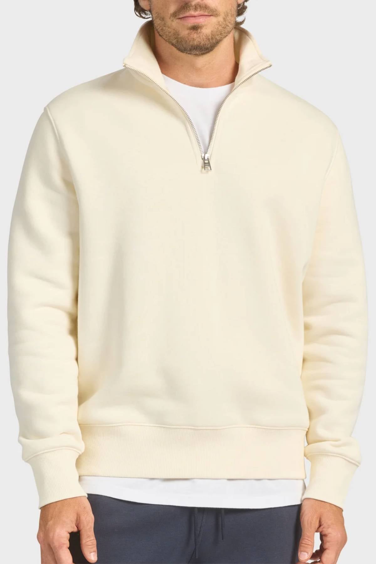 A man wears the Academy Brand 1/4 Zip Sweat in cream-colored premium brushed fleece over a white T-shirt, zipper partially open. He stands with hands by his sides in dark pants, face partly out of frame for a vintage look.