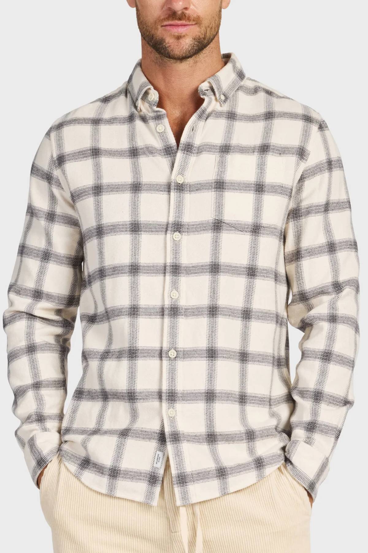 A man stands with his hands in the pockets of light-colored pants, wearing the Academy Brand Buhler Shirt—a cream shirt with a gray plaid pattern, yarn dye twill fabric, workwear buttons, a collar, and long sleeves.