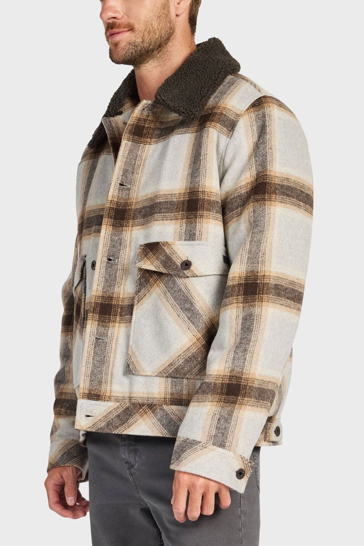 A man in the Academy Brand Conrad Jacket—a plaid wool-blend jacket in beige, brown, and gray with a dark sherpa collar and large front pockets—stands at an angle with his left hand relaxed by his side.