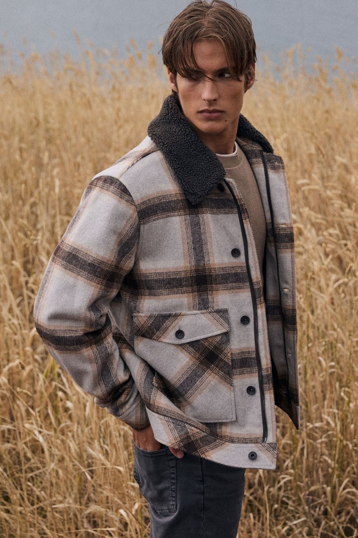 A young man stands in tall dry grass, wearing the Academy Brand Conrad Jacket—a lightweight plaid wool blend jacket featuring a dark collar—paired with a beige sweater and dark pants, looking to the side with a serious expression.