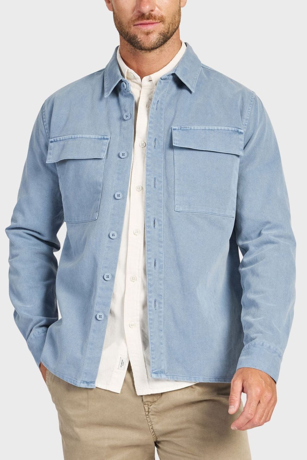 A man stands against a plain light background, wearing the Academy Brand Essential Overshirt in light blue cotton twill with double chest pockets, layered over a white collared shirt and paired with khaki pants.