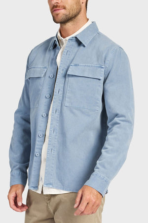 A man wears the Academy Brand Essential Overshirt in light blue cotton twill with chest pockets, layered over a white shirt and beige pants, standing against a plain light background.