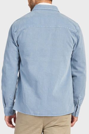 A man is seen from behind wearing the Academy Brand Essential Overshirt in light blue cotton twill with double chest pockets and beige pants, set against a plain light background.