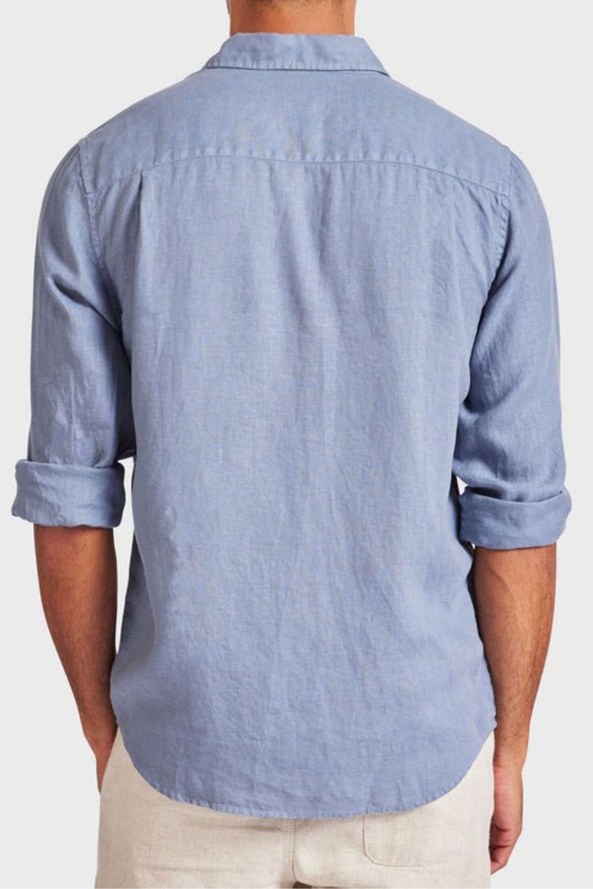 Back view of a person wearing the Academy Brand Hampton Linen Shirt in light blue, with long sleeves slightly rolled up. This lightweight staple is untucked and matched with light-colored pants against a plain background.