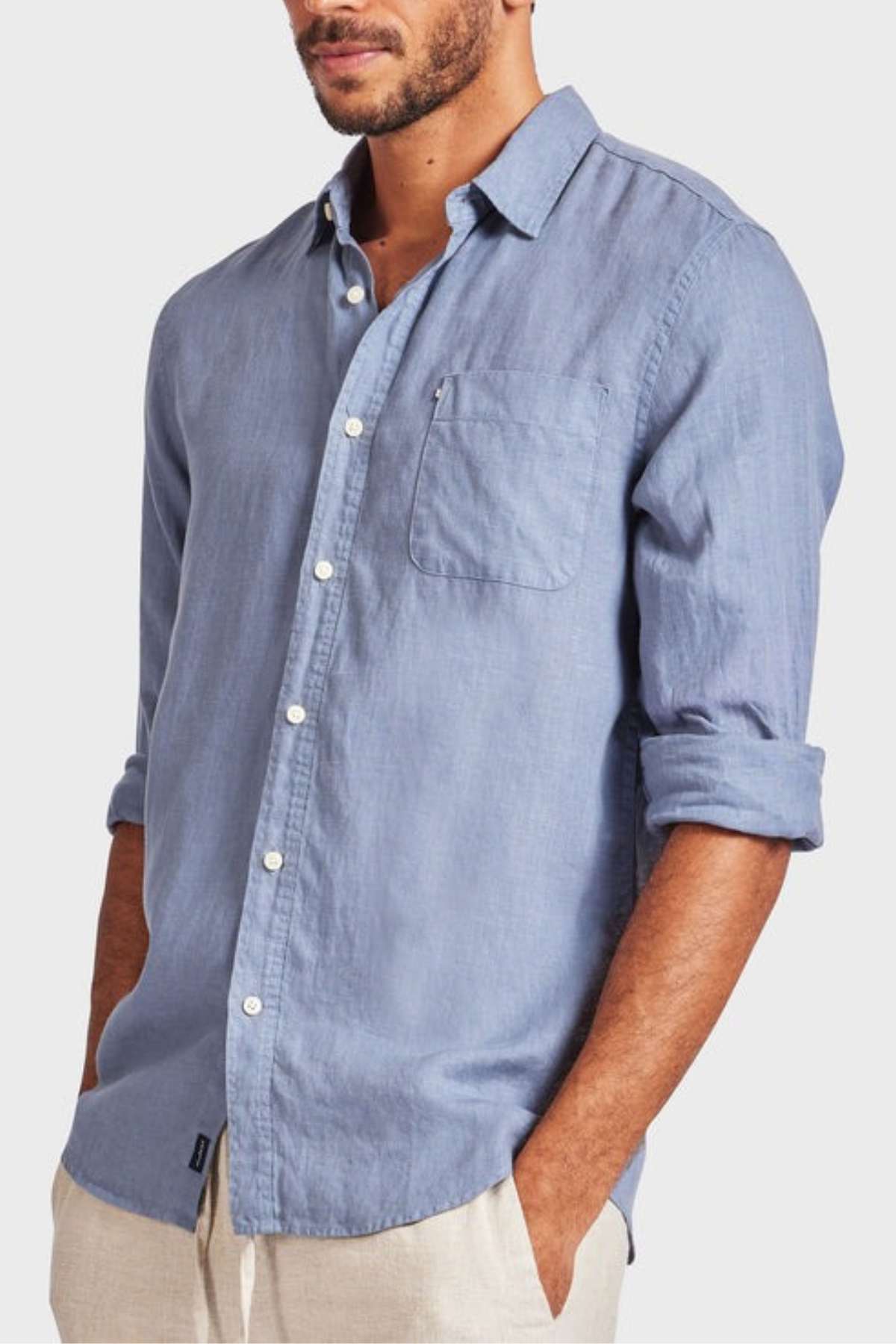 A man wearing an Academy Brand Hampton Linen Shirt in light blue, featuring rolled-up sleeves and a single chest pocket, paired with beige pants. The background is plain white.