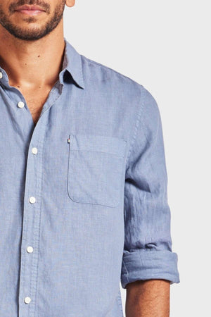 A man wearing the Academy Brand Hampton Linen Shirt in light blue is partially visible, highlighting its rolled-up sleeves for a casual vibe. This lightweight staple features a front pocket and white buttons against a plain, light-colored background.