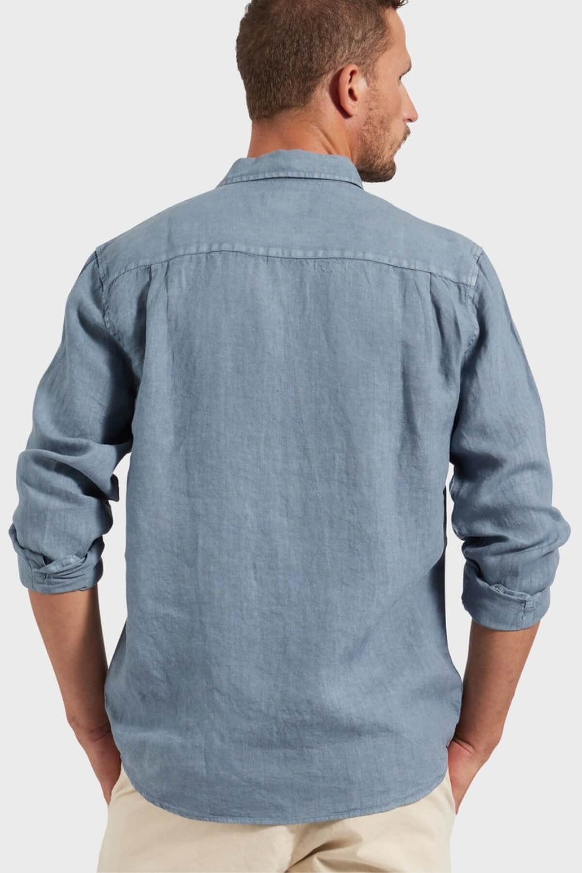 A man, seen from behind, wears a light blue Academy Brand Hampton Linen Shirt with rolled sleeves and off-white pants, standing against a plain light gray background.