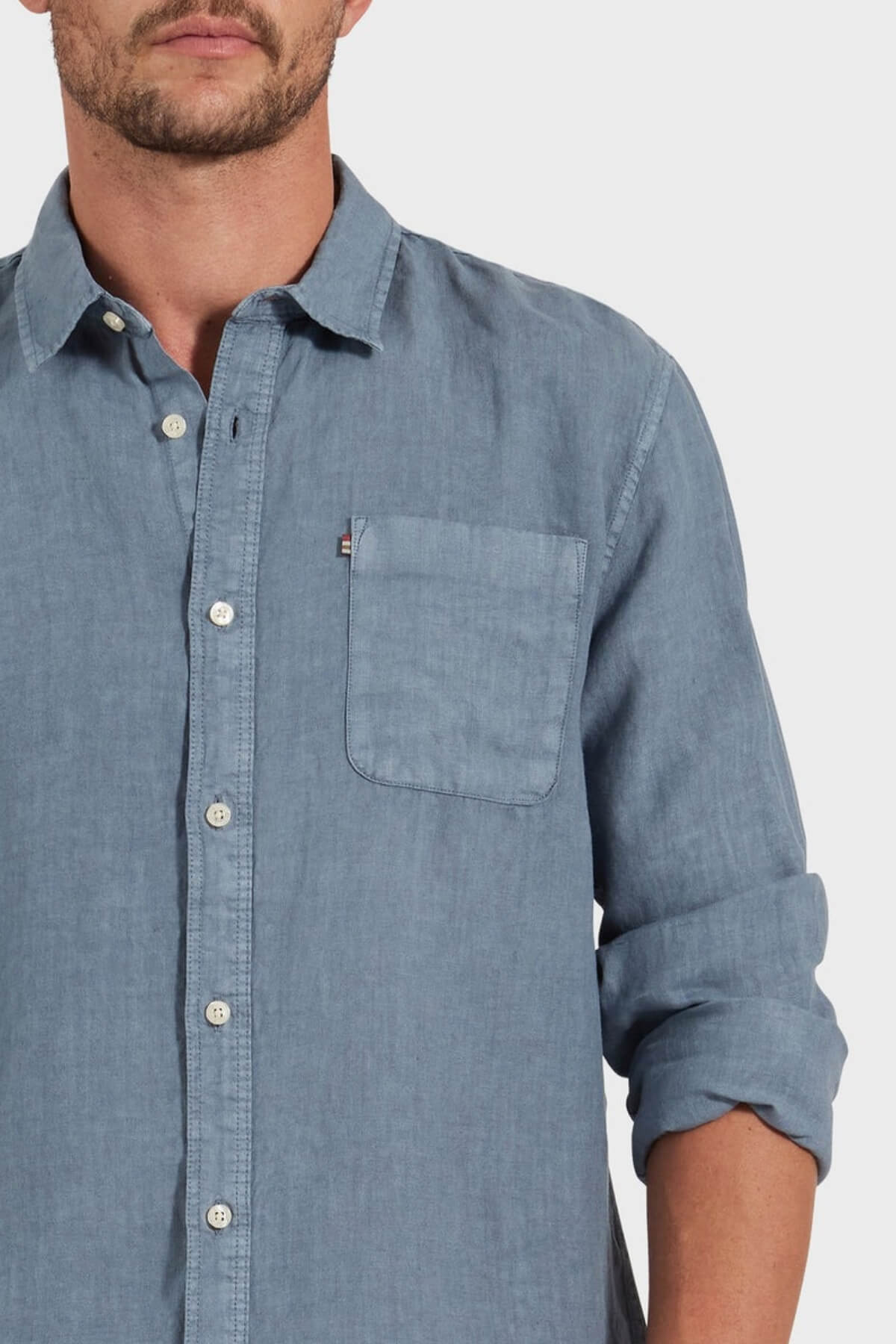 A man wears an Academy Brand Hampton Linen Shirt in light blue with a left chest pocket and rolled-up sleeves. His face is partially out of frame against a plain white background.