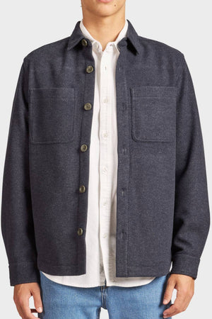 A person wears the Academy Brand Lloyd Overshirt, a dark gray men's overshirt with two chest pockets, styled over a white shirt and blue jeans. The jacket is partly unbuttoned and the person's face is not shown.