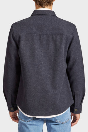 A man is seen from behind wearing the Academy Brand Lloyd Overshirt in dark gray, layered over a white shirt and light blue jeans. The photo focuses on the back and sleeve features of the overshirt.