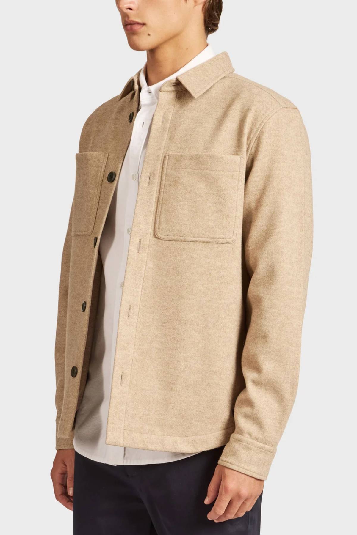 A person wears the Academy Brand Lloyd Overshirt, a beige button-up men’s shirt with two chest pockets, layered over a white shirt and dark pants, standing before a plain light background.