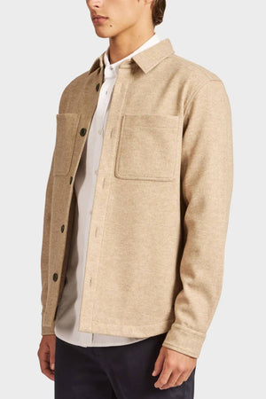 A person wears the Academy Brand Lloyd Overshirt, a beige button-up men’s shirt with two chest pockets, layered over a white shirt and dark pants, standing before a plain light background.