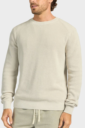 A man wears the Academy Brand Oliver Knit Crew, a light beige long-sleeve sweater made from cotton yarn, paired with matching beige drawstring pants. He stands against a plain background with his face partially visible.