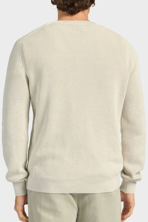 A person is seen from behind wearing the Academy Brand Oliver Knit Crew, a light beige, relaxed-fit sweater made from soft cotton yarn with a vintage wash and ribbed crew neckline. They pair it with light-colored pants against a plain white background.