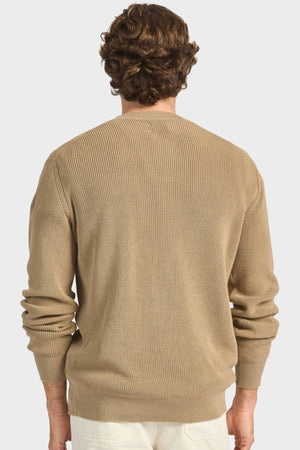 A man with wavy brown hair is shown from the back, wearing the Academy Brand Oliver Knit Crew in ribbed premium cotton yarn and white pants against a plain light background.