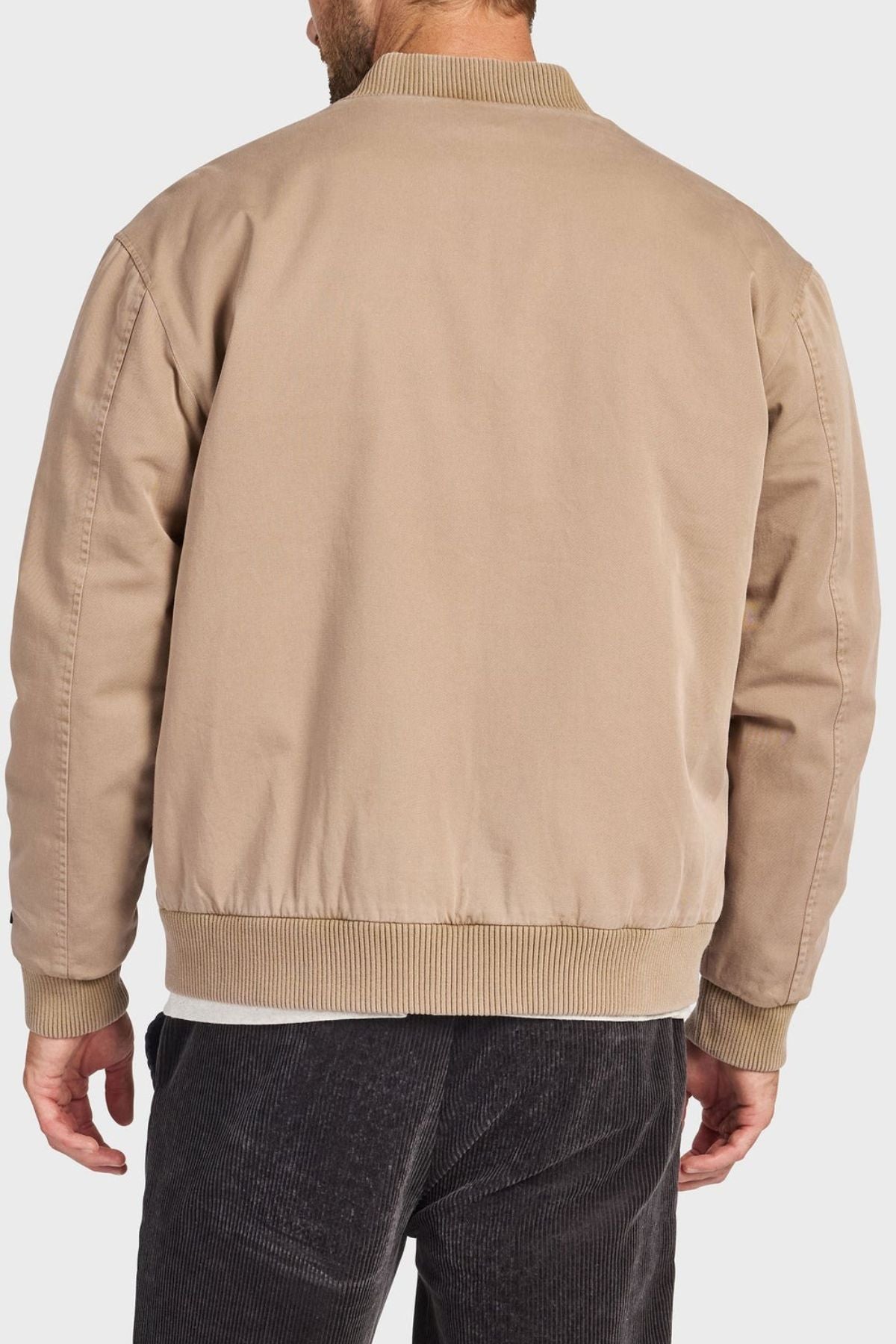 A man is seen from the back wearing the Academy Brand Standard Issue Bomber, a light brown cotton twill jacket with ribbed cuffs and hem, paired with dark pants.