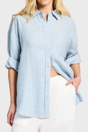 A person wearing a light blue Academy Women Hampton Linen Shirt, complete with rolled-up sleeves and mother-of-pearl buttons, pairs it effortlessly with white pants. With one hand resting in their pocket against a neutral background, they exude a relaxed yet polished vibe.