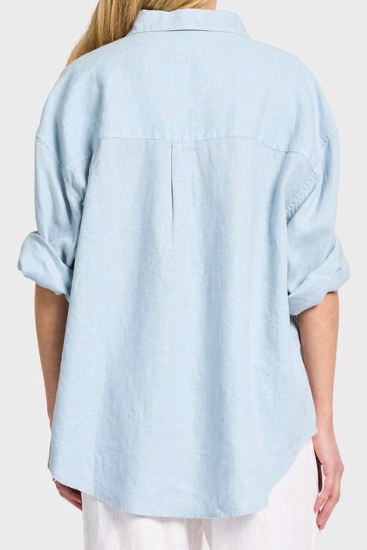 A person is seen from the back wearing an Academy Women Hampton Linen Shirt in light blue with rolled-up sleeves and mother-of-pearl buttons. The garment-dyed shirt has a loose fit that perfectly complements their long blond hair against a plain gray background.