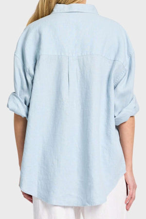 A person is seen from the back wearing an Academy Women Hampton Linen Shirt in light blue with rolled-up sleeves and mother-of-pearl buttons. The garment-dyed shirt has a loose fit that perfectly complements their long blond hair against a plain gray background.