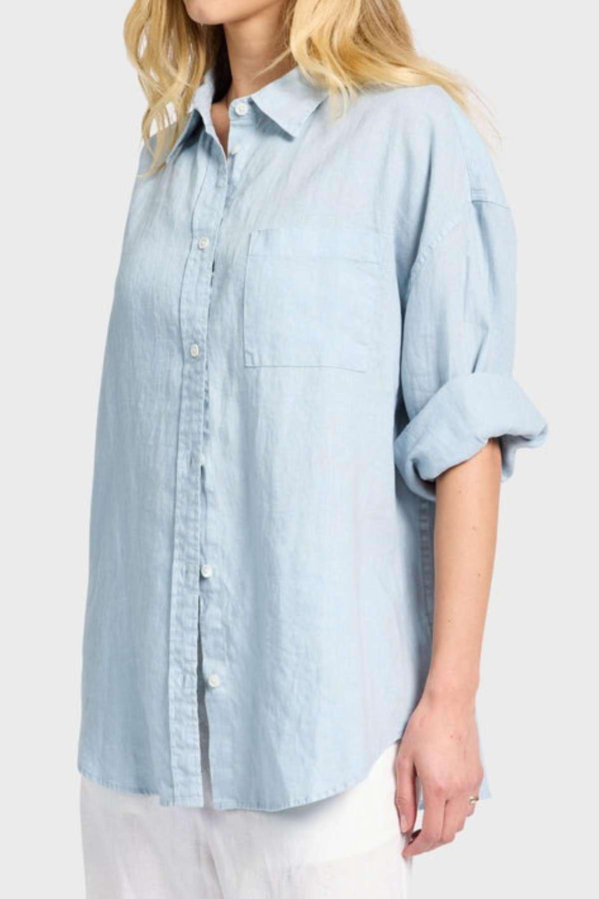 A person donning a light blue Academy Women Hampton Linen Shirt, accentuated with mother-of-pearl buttons, rolled-up sleeves, and a pocket on the left chest, is paired with white pants against a plain background. The individual's face is not visible.