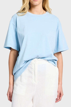 A person is wearing a light blue Academy Women Jimmy Boyfriend Tee, made from premium sanded jersey, tucked into high-waisted white pants. They stand against a plain gray background with long blonde hair cascading down, their hands resting at their sides. The outfit exudes a casual and relaxed vibe.