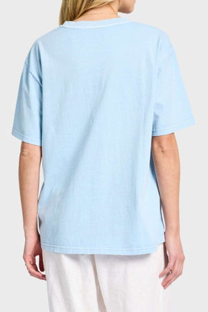 A person with light hair is wearing an oversized, light blue Academy Women Jimmy Boyfriend Tee and white pants, standing with their back to the camera against a plain gray background.
