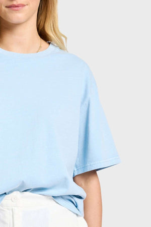 A person is wearing an Academy Women Jimmy Boyfriend Tee in a sanded jersey fabric, paired with white pants. Only their upper torso is visible against a plain gray background, complementing their long, light-colored hair.