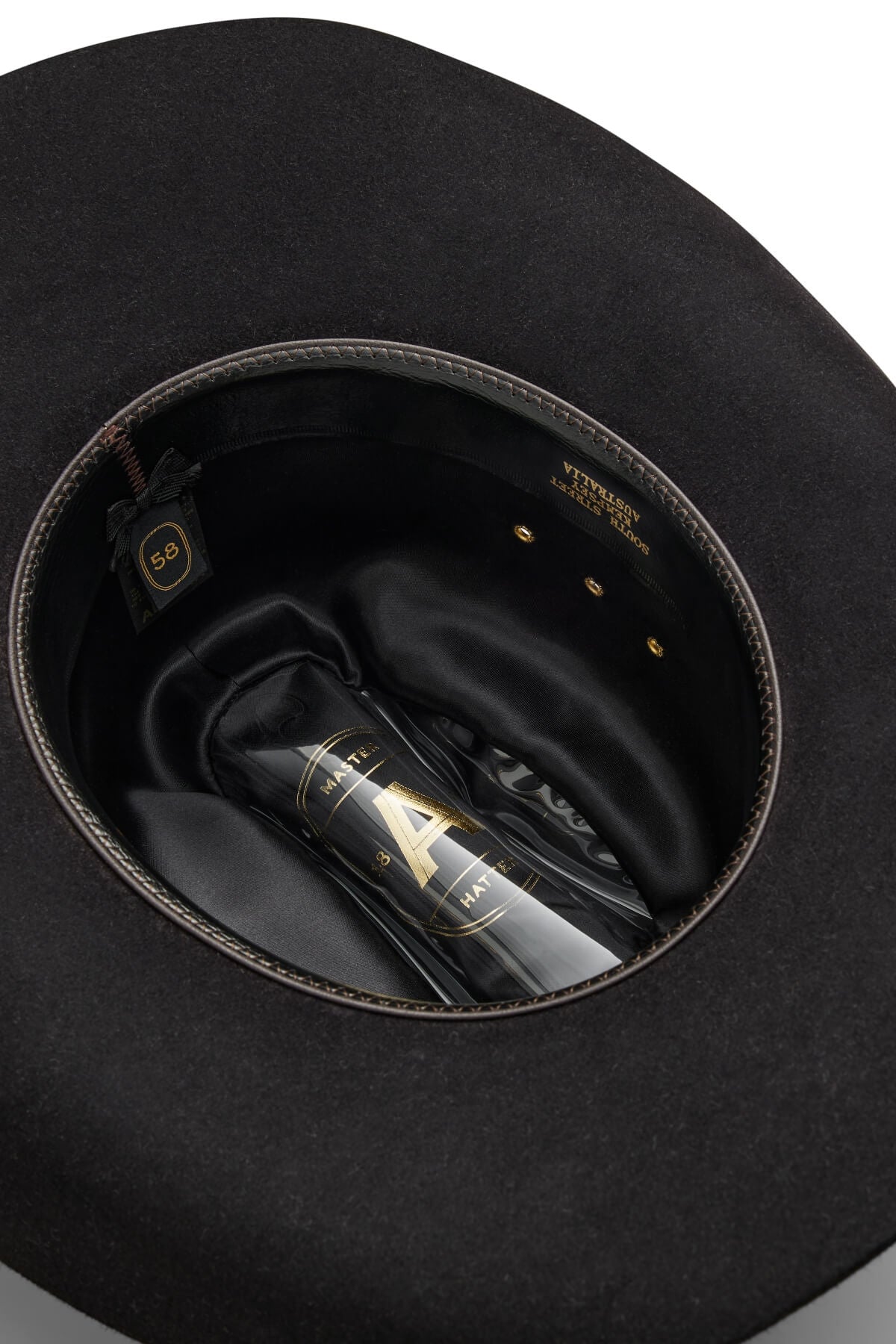 An Akubra Outback Club hat in black, size 58, sits upside down to display its inner lining and a clear bottle with gold and black labeling nestled in the crown—a tribute to Lee Kernaghan and Australian country music.