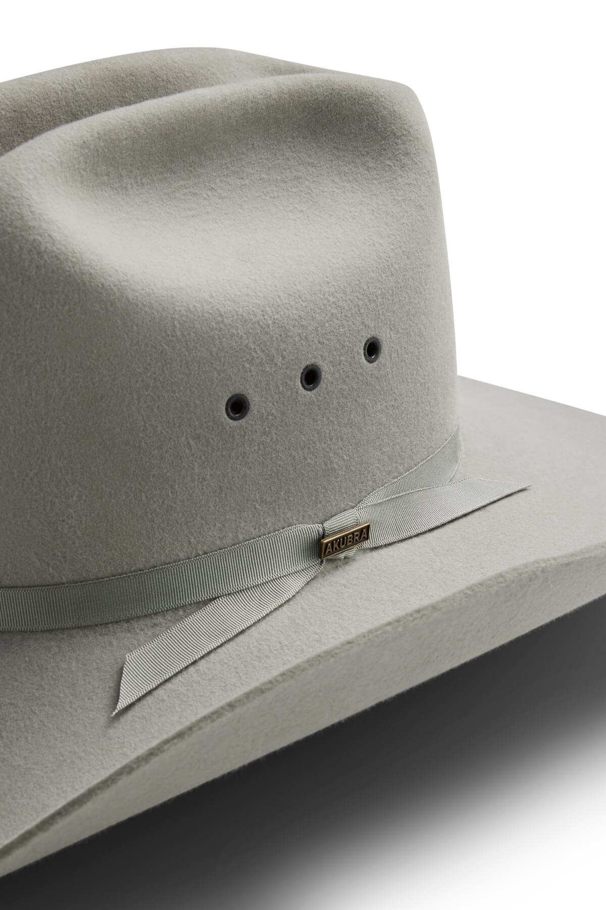 A close-up of a light grey Akubra Outback Club hat features a matching ribbon band, three ventilation holes, and a small metal AKUBRA tag on the ribbon. Inspired by Australian country music legends. White background.