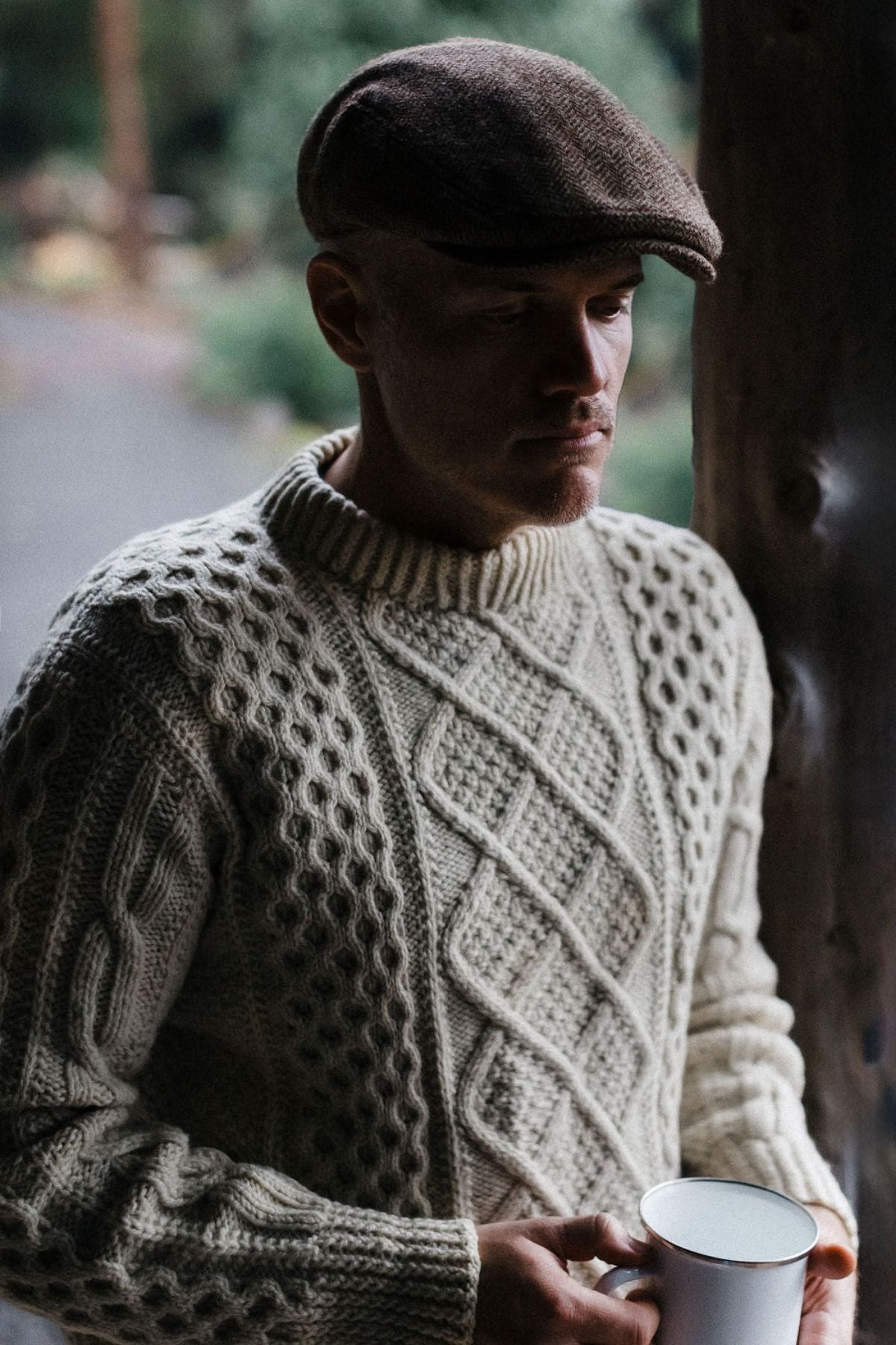 A man in a textured cream sweater and the Avenel Cunningham Six Panel Nailhead Wool Blend Cap stands by a wooden post, holding a white cup and looking down thoughtfully, with blurred greenery in the background.