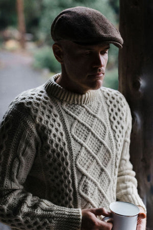 A man in a textured cream sweater and the Avenel Cunningham Six Panel Nailhead Wool Blend Cap stands by a wooden post, holding a white cup and looking down thoughtfully, with blurred greenery in the background.