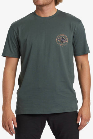 A person with light skin and shoulder-length light brown hair is wearing a dark green Billabong Rotar Diamond T-Shirt, which has a small geometric logo pattern on the left chest area. The regular fit tee is made of 100% cotton. They are also dressed in black pants or shorts.