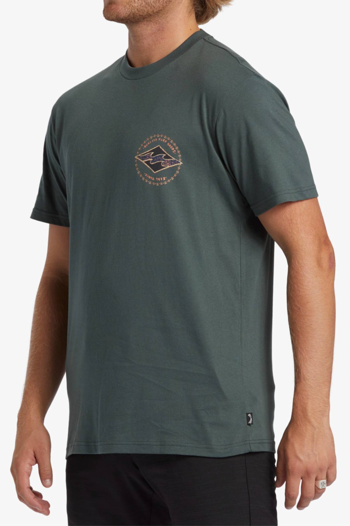 A man is wearing a Billabong Rotar Diamond T-Shirt, a dark green, short-sleeved tee with a small circular logo on the left chest area. Made of 100% cotton, this regular fit shirt features a mountain and text around the logo. He has short, light brown hair and is also dressed in black pants.