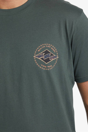 A person is wearing a dark green Billabong Rotar Diamond T-Shirt, made of 100% cotton with a small emblem on the left chest. The emblem features a blue academic cap encircled by the words "MAN IITIVI EVRE GODS" and "SINCE 1317," along with a decorative border. This regular fit T-shirt effortlessly combines style and comfort.