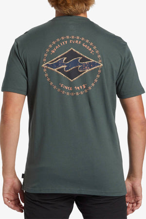 A person with light brown hair is wearing a Billabong Rotar Diamond T-Shirt in dark green. The back of this regular fit T-shirt showcases a circular logo featuring the text "Quality Surf Goods" at the top and "Since 1973" at the bottom, framing a stylized wave graphic in the center.