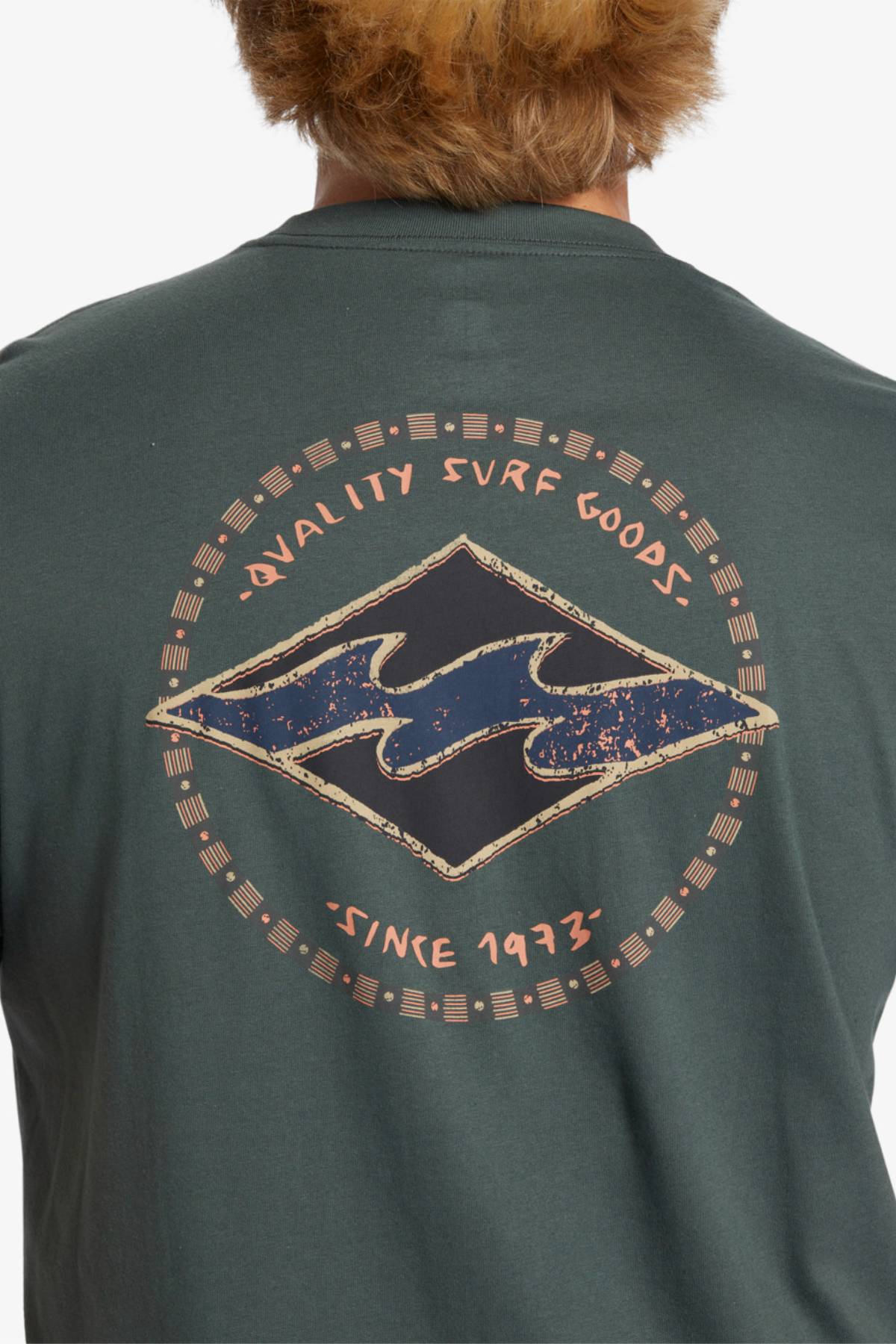 A person with light brown hair is wearing a dark green Billabong Rotar Diamond T-Shirt. The regular fit shirt features a circular design on the back, with "QUALITY SURF GOODS" at the top and "SINCE 1973" at the bottom, surrounding a blue wave symbol.