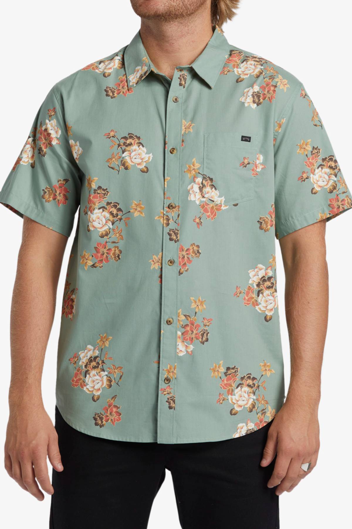 A man wears the Billabong Sundays Mini Short Sleeve Shirt, a light green shirt with bold orange, white, and yellow floral prints. He stands with his arms relaxed at his sides.