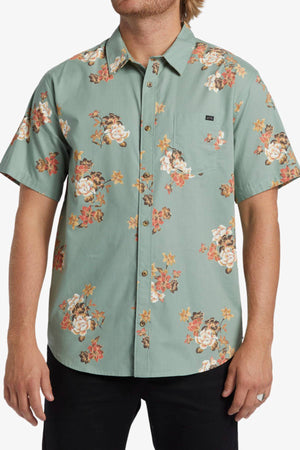 A man wears the Billabong Sundays Mini Short Sleeve Shirt, a light green shirt with bold orange, white, and yellow floral prints. He stands with his arms relaxed at his sides.