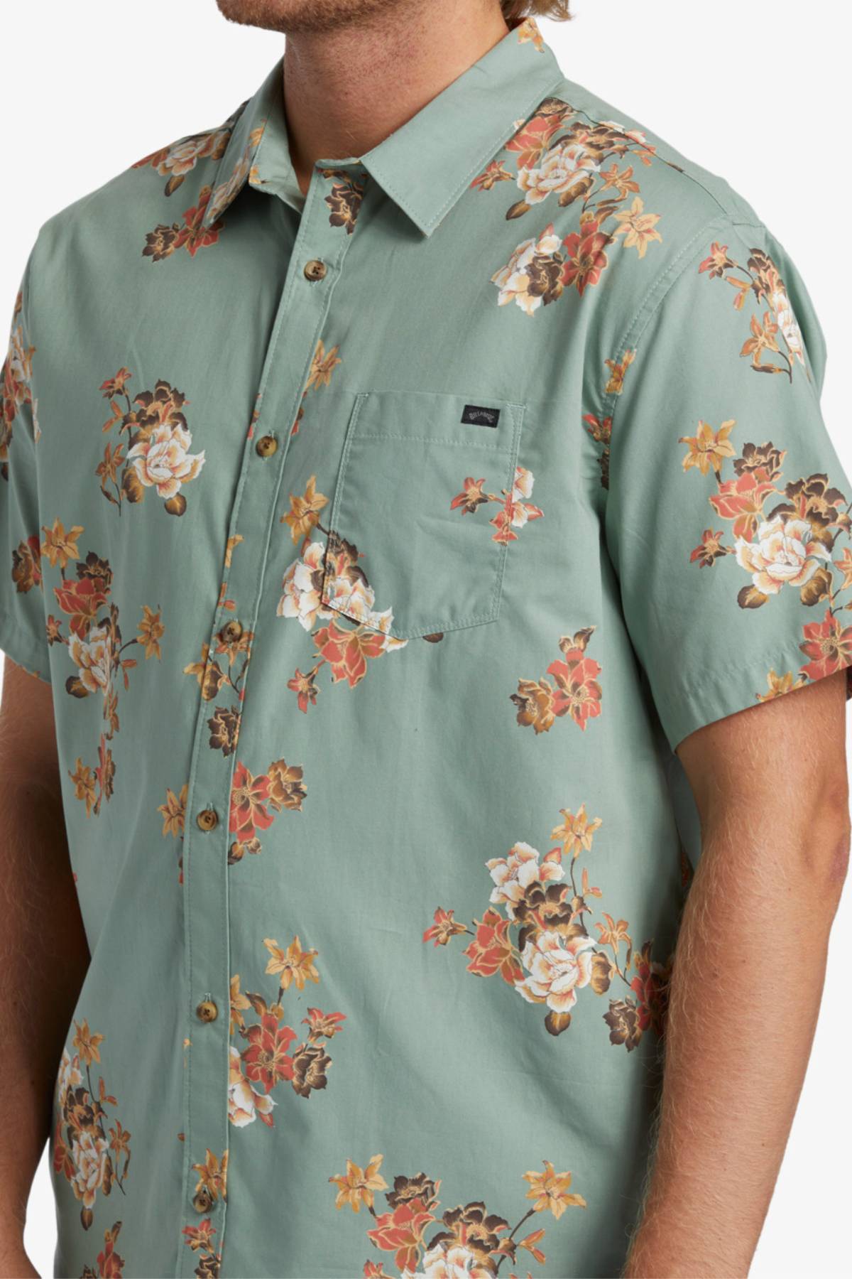 A man wears the Billabong Sundays Mini Short Sleeve Shirt, a light green short sleeve top with a bold white, orange, and brown floral print, featuring a chest pocket accented by a small black label.