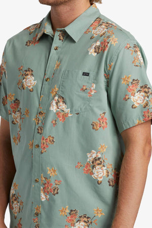 A man wears the Billabong Sundays Mini Short Sleeve Shirt, a light green short sleeve top with a bold white, orange, and brown floral print, featuring a chest pocket accented by a small black label.