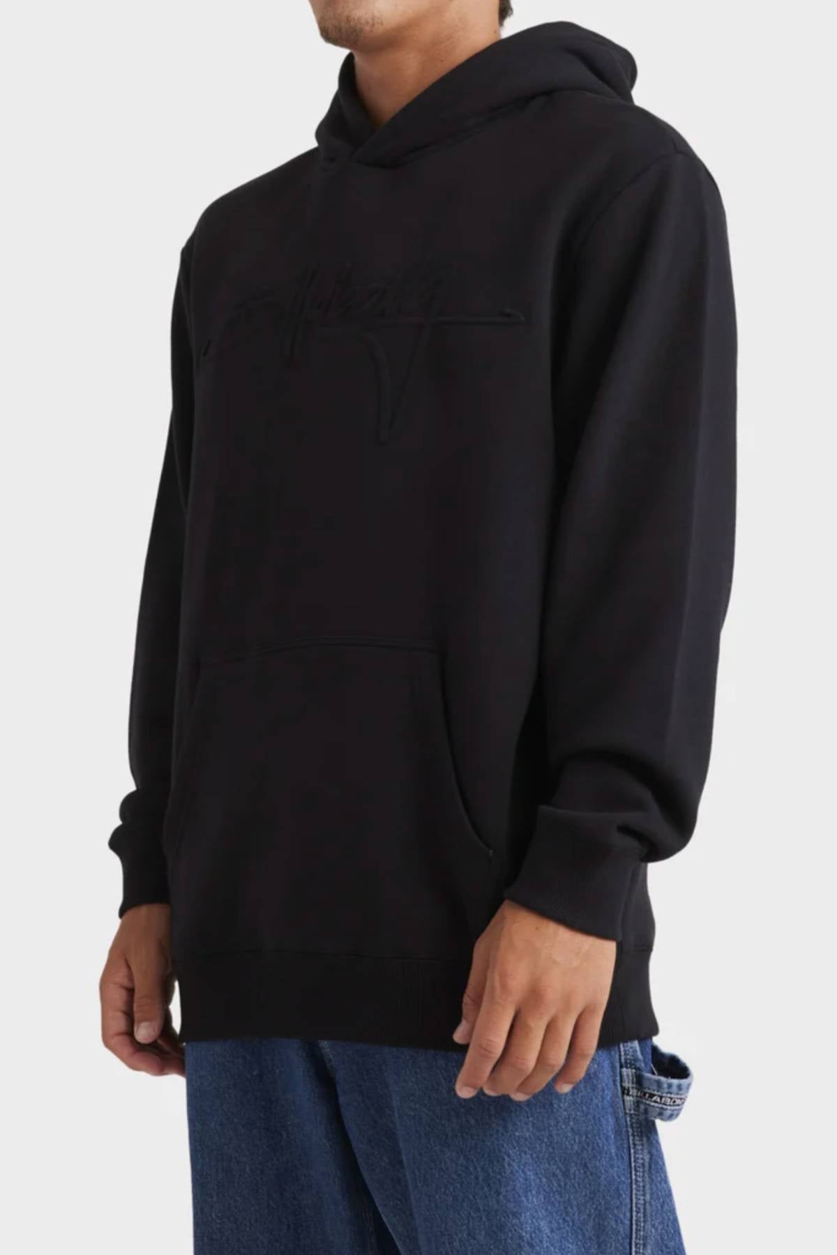 A person wears the Billabong Tonal Boss Pop Hood, a black fleece sweatshirt with subtle stitched chest lettering, paired with blue jeans and standing against a plain light background.