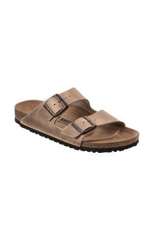 A single Birkenstock Arizona sandal in tan, featuring two adjustable buckle straps and a cork footbed. The sandal has a black rubber sole and an inscription on the side of the footbed.