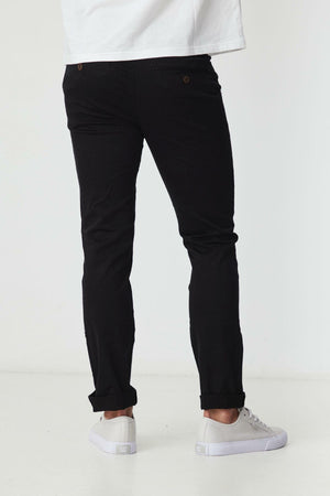 A person stands facing away, wearing BLKWD Clay Slim Chino Pant in black, made from cotton stretch fabric, paired with a white shirt and white sneakers against a plain white background.