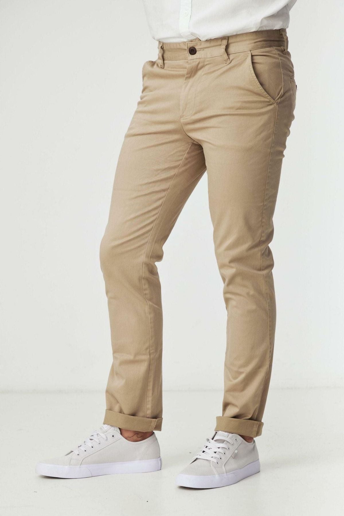 A person wears BLKWD Clay Slim Chino Pant in beige with a white shirt and white sneakers, standing against a plain white background. The person's head is not visible in the image.