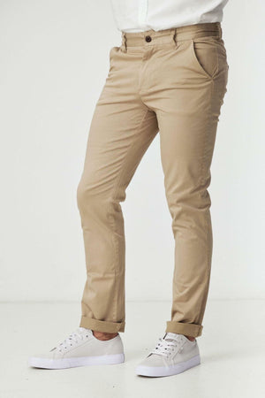 A person wears BLKWD Clay Slim Chino Pant in beige with a white shirt and white sneakers, standing against a plain white background. The person's head is not visible in the image.